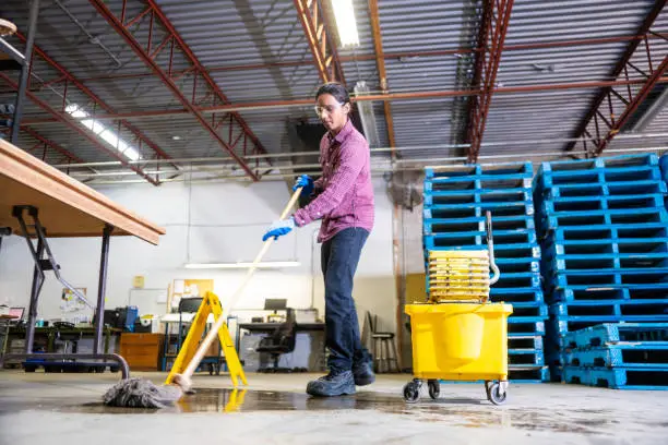 Professional warehouse cleaning in Cambridge MA featuring an industrial cleaner mopping a concrete facility floor near blue shipping pallets and safety equipment.